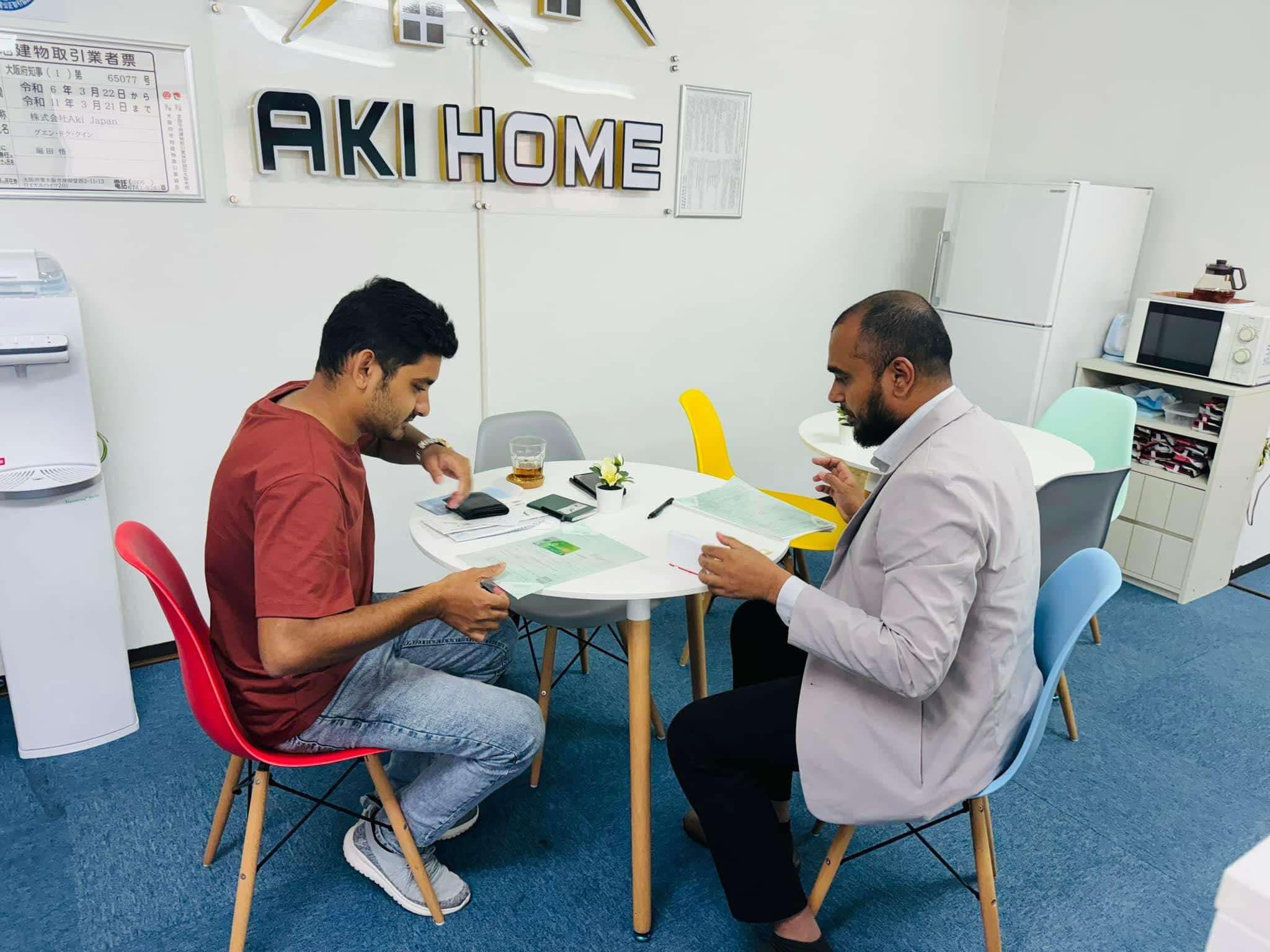 Two clients reviewing paperwork with a consultant under the AKI HOME office sign — paid job placement support